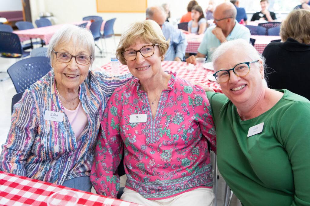 Three individuals wearing name tags smile for the camera while seated at a table with a red and white checkered tablecloth in a social gathering setting.